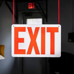 May include: A white sign with red lettering that says "EXIT" hanging from the ceiling.