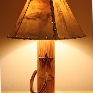 May include: Rustic wooden table lamp with a brown leather lampshade. The lamp features a wooden base, a metal star, and a horseshoe. The lampshade is stitched with dark thread. The lamp is designed in a western style.