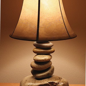 May include: A table lamp with a beige fabric shade and a base made of stacked stones. The lamp is lit and casting a warm glow.