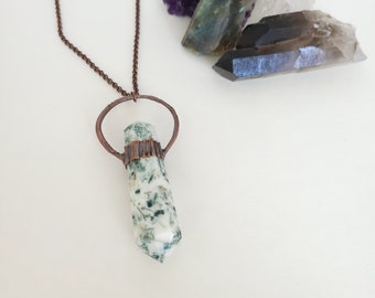 May include: A copper chain necklace with a green and white agate pendant. The pendant is wrapped in copper wire and has a copper ring at the top. The necklace is displayed on a white surface with various crystals.
