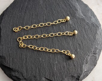 Puede incluir: Tres cadenas doradas con extremos de bola redonda. Las cadenas están sobre una superficie negra.