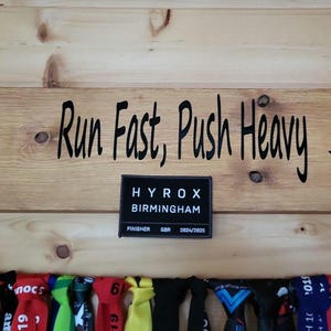 May include: A wooden wall with the text "Run Fast, Push Heavy" and a black and white logo for Hyrox Birmingham. Below the text are several colourful medal ribbons.