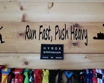 Könnte beinhalten: Eine Holzwand mit dem Text "Run Fast, Push Heavy" und einem schwarz-weißen Logo für Hyrox Birmingham. Unter dem Text befinden sich mehrere bunte Medaillenbänder.