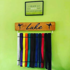 K&ouml;nnte beinhalten: Ein Holz-G&uuml;rtelhalter mit dem eingravierten Namen "Luke". Der Halter hat an jedem Ende eine Silhouette einer Person, die einen Karate-Tritt ausf&uuml;hrt. Es h&auml;ngen 12 G&uuml;rtel am Halter, darunter wei&szlig;e, lila, gelbe, orange, gr&uuml;ne, braune und schwarze G&uuml;rtel.