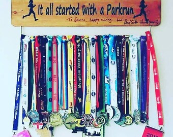 Könnte beinhalten: Ein Holzschild mit dem Text "It all started with a Parkrun" und einer laufenden Figur auf jeder Seite. Das Schild hängt an einer Wand mit einem Metallgestell, das eine Sammlung bunter Laufmedaillen und Bänder hält.