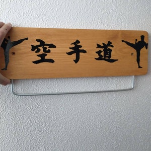 K&ouml;nnte beinhalten: Ein Holzschild mit den japanischen Schriftzeichen "空手道" (Karate-do) in schwarzer Farbe. Zwei schwarze Silhouetten von Karate-Praktizierenden befinden sich auf beiden Seiten des Textes.