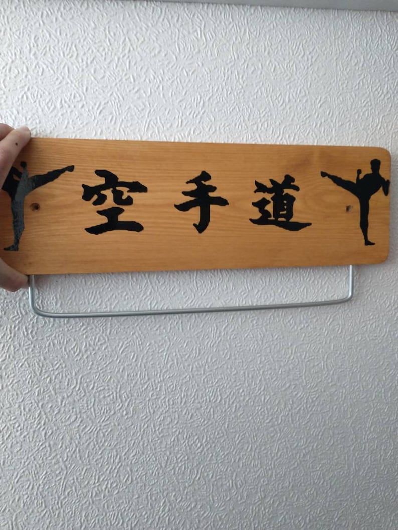K&ouml;nnte beinhalten: Ein Holzschild mit den japanischen Schriftzeichen "空手道" (Karate-do) in schwarzer Farbe. Zwei schwarze Silhouetten von Karate-Praktizierenden befinden sich auf beiden Seiten des Textes.