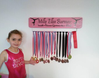 Könnte beinhalten: Ein rosafarbenes Holz-Medaillenbrett mit einer Turner-Silhouette und dem Text "Milly Elba Burman South Devon Gymnastics Club". Das Brett hat einen silbernen Metallhaken zum Aufhängen von Medaillen. Ein junges Mädchen in einem rosafarbenen Trikot mit dem Text "South Devon" steht vor dem Brett.