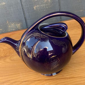 Puede incluir: Una tetera azul cobalto con ribete dorado y un diseño floral. La tetera tiene un cuerpo redondeado y un asa curva.