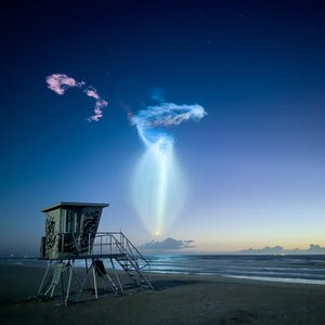 May include: A nighttime beach scene featuring a lifeguard tower and a bright, glowing light formation in the sky. The tower is gray with graffiti, and the sky transitions from dark blue to lighter hues. The ocean and sand are visible in the foreground.