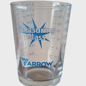 May include: Clear glass measuring cup with blue text and markings. The cup has a starburst design with the words "Measuring Glass" and the brand "ARROW". Graduated measurements in ounces and milliliters are visible.