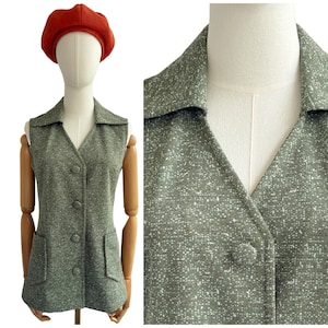 Könnte beinhalten: Eine olivgrüne Vintage-Weste mit gesprenkeltem Muster, Kragen, Knopfleiste und zwei Vordertaschen. Die Weste wird auf einer Schaufensterpuppe mit roter Baskenmütze präsentiert.