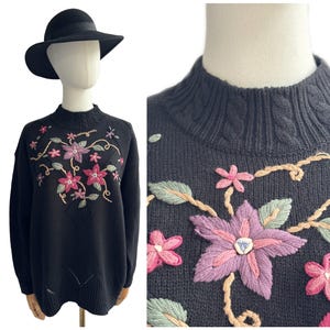 May include: Black sweater with embroidered floral design in pink, purple, and green. The sweater has a mock neck and long sleeves. A black hat is on the mannequin's head. The sweater's floral embroidery is the focal point.