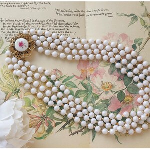 May include: A vintage necklace with multiple strands of white beads and gold-coloured spacers. The necklace has a decorative gold-coloured clasp with a white centre and a pink floral design. The necklace is displayed on a floral background.