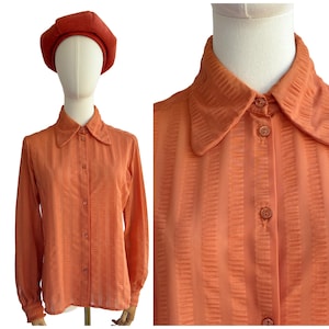 May include: An orange vintage button-down shirt with vertical textured stripes, long sleeves, and a pointed collar. The shirt is displayed on a mannequin and paired with a matching orange beret.