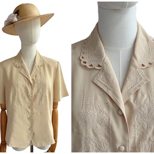 May include: A short-sleeved beige button-down blouse featuring intricate tonal floral embroidery on the front panels and a scalloped collar. The blouse is displayed on a mannequin wearing a matching beige hat with a white floral accent.