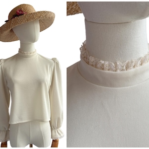May include: Cream-coloured long-sleeve blouse with a high, ruffled collar and cuffs. The blouse has a slight puff sleeve design. A straw hat with a pink flower is on top of the mannequin head.