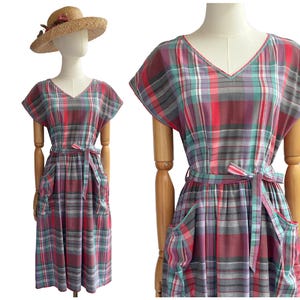 May include: A vintage plaid dress in shades of red, gray, and teal, displayed on a mannequin. The dress features a V-neck, short sleeves, a matching belt, and pockets. A straw hat with a floral accent sits atop the mannequin's head, completing the ensemble.