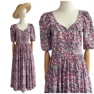 Peut inclure: Robe vintage à imprimé floral avec un décolleté en V et des manches courtes. La robe présente un fond sombre avec des motifs floraux roses et verts. Un chapeau de paille est sur une tête de mannequin. La robe a un corsage ajusté et une jupe fluide.