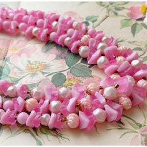 May include: A vintage pink necklace with multiple strands. The necklace features a combination of pink beads, irregular-shaped pink pieces, and white pearls. The necklace is displayed on a floral patterned background.