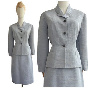 Könnte beinhalten: Hellblauer Vintage-Rockanzug. Die Jacke hat einen Reverskragen und drei dunkle Knöpfe. Der Rock ist knielang. Der Anzug wird auf einer Schaufensterpuppe präsentiert. Der Stoff hat eine strukturierte Optik.