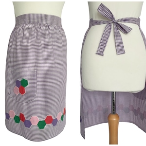 May include: A purple and white gingham apron with a pocket and a decorative floral design made of colorful hexagons. The apron ties at the waist with a matching gingham bow.