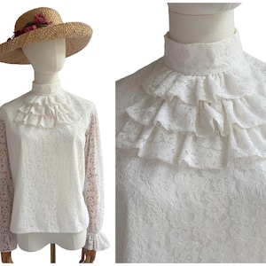May include: A white long-sleeved blouse featuring a high neck, lace fabric, and a multi-layered ruffled bib front. The blouse is displayed on a mannequin wearing a straw hat adorned with small pink flowers and green leaves.