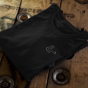Op de afbeelding: Een zwart T-shirt met een wit slangontwerp geborduurd op de voorkant.