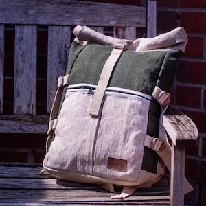 May include: A green and beige canvas rucksack with a roll-top closure and adjustable straps. The rucksack has a zipped pocket on the front and a large main compartment.