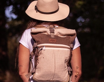 May include: A person wearing a brown and beige canvas backpack with a brown leather strap and a brown and beige roll-top closure. The backpack has a brown leather patch with the word "Adventure" on it. The person is also wearing a brown straw hat.