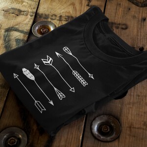 Peut inclure: T-shirt noir avec un motif de flèches blanches. Les flèches présentent divers éléments décoratifs, notamment des plumes, des formes géométriques et un motif en forme de blé. Le t-shirt est plié sur une surface en bois.