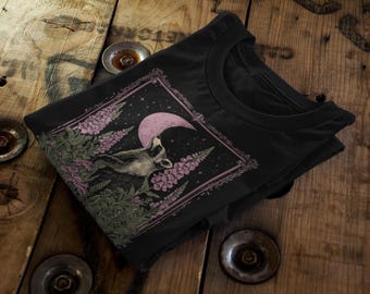 May include: Black t-shirt featuring a raccoon howling at a pink crescent moon, surrounded by purple flowers and green foliage. The design is framed with a decorative border. The shirt is folded on a wooden surface.