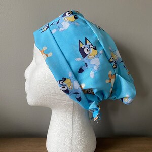May include: A light blue surgical scrub cap featuring a repeating pattern of a cartoon dog character in various poses. The cap is tied at the back and is displayed on a white foam head.