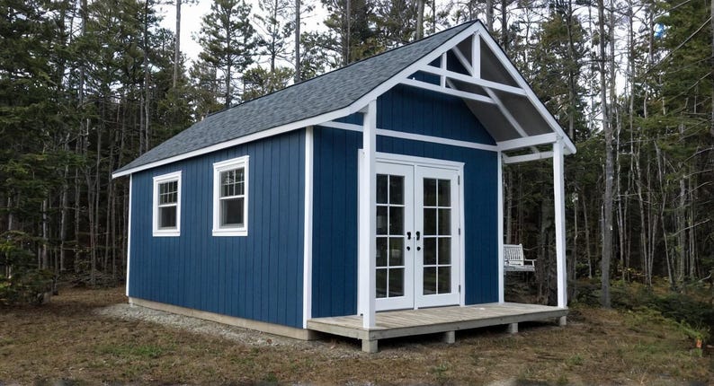 A blue and white cabin in the woods with a beautiful front porch and gable roof.