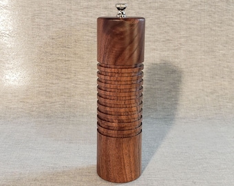 Op de afbeelding: Een cilindrische houten pepermolen met een zilveren bovenkant. De molen is gemaakt van donker hout met een gelaagd ontwerp.