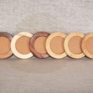 May include: Six circular coasters with cork centres and wooden frames. The wood frames vary in colour, from dark brown to light oak. The coasters are arranged in a row, showcasing the different wood tones.