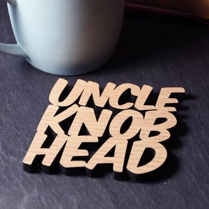 May include: A pale blue mug next to a wooden coaster with the words "UNCLE KNOB HEAD." A globe and an open book with flowers are in the background. The coaster is made of light-coloured wood.