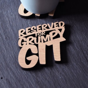 Può includere: Un sottobicchiere di legno con il testo "Riservato per Grumpy Grrrl" in un font stilizzato. Il sottobicchiere è di colore marrone chiaro e ha una superficie liscia. Una tazza blu chiaro è appoggiata sul sottobicchiere.