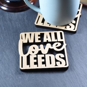 May include: Two wooden coasters with the text "We All Love Leeds" on one coaster. The coasters are on a black surface with a blue mug and a globe in the background.