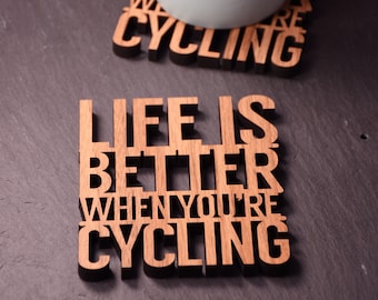 May include: Two wooden coasters with the text "Life is better when you're cycling" and "We are cycling". A light blue mug is sitting on top of the "We are cycling" coaster.