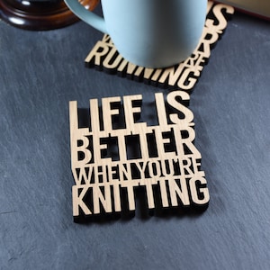 May include: A light blue mug sits on a wooden coaster that reads "Life is better when you're knitting". The coaster is on a dark gray surface. A globe and a book are in the background.