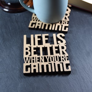 May include: Two wooden coasters with the text "Life is better when you're gaming" and "When you're gaming" in black lettering. A light blue mug sits on top of the coasters.