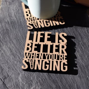 May include: Two wooden coasters with a light brown finish. The top coaster has the text "Life is better when you're singing" with a microphone icon. The bottom coaster has the text "Better when you're singing" with a microphone icon.
