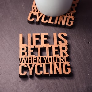 Może przedstawiać: Dwa drewniane podkładki z tekstem "Life is better when you're cycling" i "We are cycling". Jasnoniebieski kubek stoi na podkładce "We are cycling".
