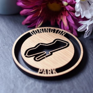Könnte beinhalten: Ein Holz-Untersetzer mit einer schwarzen Umrandung der Donington Park Rennstrecke. Der Untersetzer liegt auf einer dunkelgrauen Oberfläche mit einer rosa und weißen Blume.