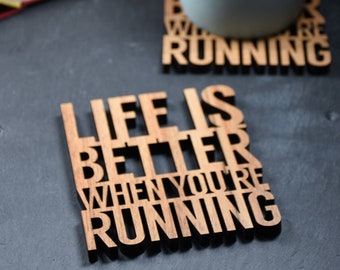 Może przedstawiać: Drewniane podkładki pod kubki z tekstem "Life is better when you're running" i "Where you're running". Podkładki znajdują się na czarnej powierzchni z niebieskim kubkiem i książką.