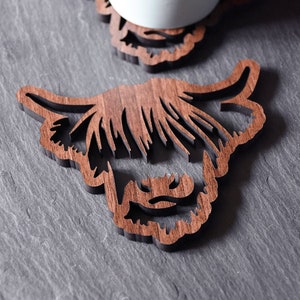May include: Two wooden coasters with a Highland cow design. One coaster supports a white mug. The coasters are made of dark brown wood and are cut in the shape of a Highland cow's head. The coasters are on a gray surface.