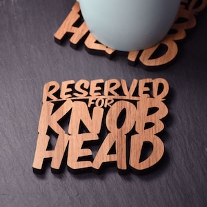 May include: Two wooden coasters with a brown wood grain finish. The coaster on the left says "Reserved for Knob Head". The coaster on the right says "Head".