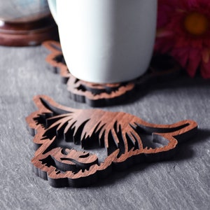 May include: A white mug rests on a wooden coaster with a detailed, layered design. The coaster is shaped like a Highland cow's head, with intricate cutouts. The background includes a globe and a pink flower, suggesting a home decor theme.