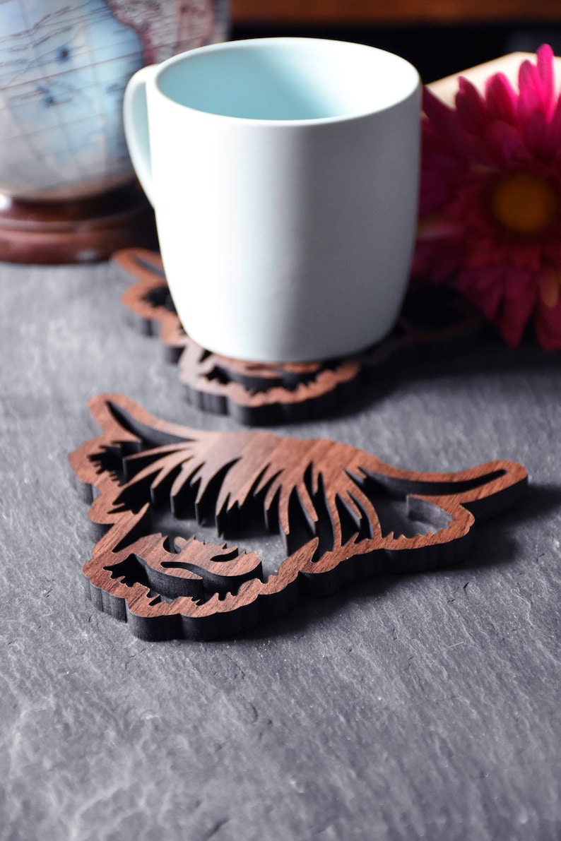 May include: A white mug rests on a wooden coaster with a detailed, layered design. The coaster is shaped like a Highland cow's head, with intricate cutouts. The background includes a globe and a pink flower, suggesting a home decor theme.